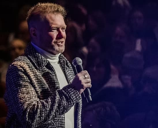 Craig Ralston zingt in een microfoon op het podium, met dramatische concertverlichting