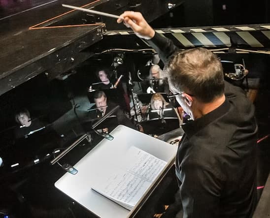 Craig Ralston dirigeert een ensemble vanaf de lessenaar, dirigeerstok omhoog, partituur open voor hem