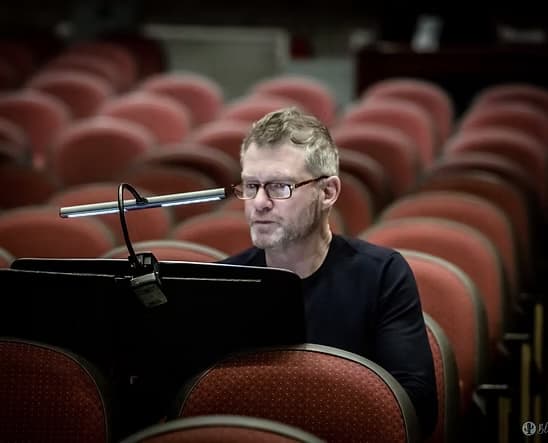 Craig Ralston seated at a conductor's stand in an empty theatre, reviewing a score